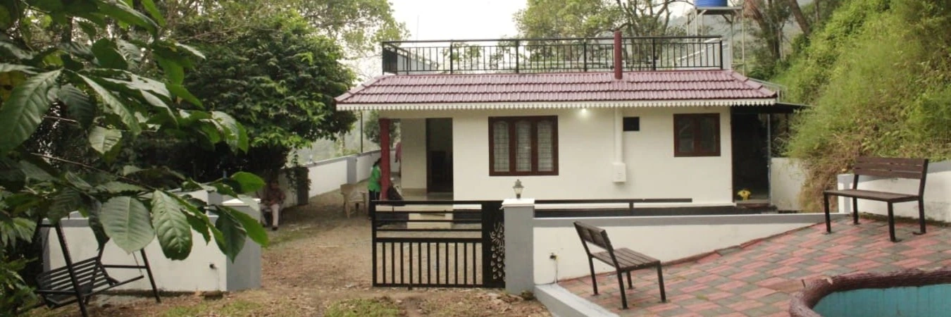 Single-story white building of Valley Homestay Munnar featuring a paved front courtyard and garden seating areas.