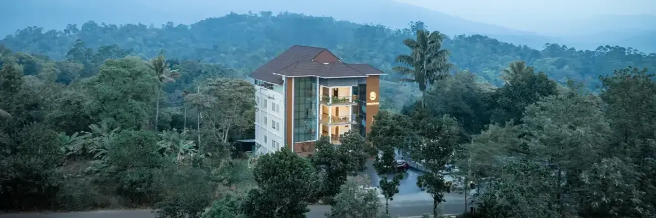 Multi-story white building of Boulevard Hotel Munnar with a dark gabled roof nestled in a dense green forest landscape