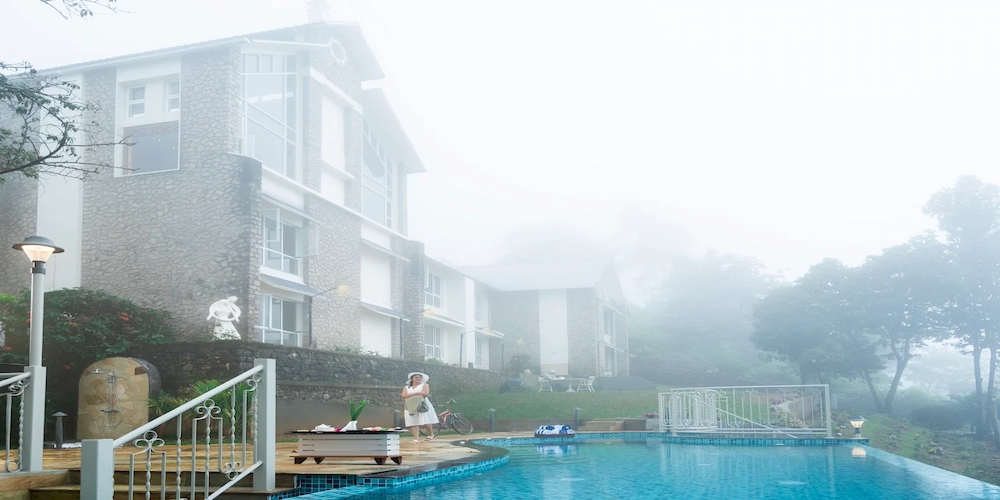Outdoor swimming pool surrounded by misty mountains at 1924 Clock House Resort Munnar