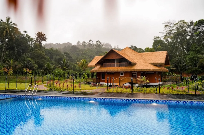 Outdoor swimming pool reflecting traditional resort building under blue sky at The Hamlet Farmstead Munnar